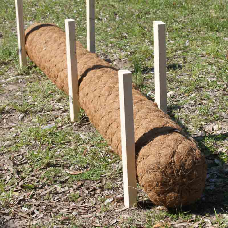 Close-up of a coir log secured by wooden stakes for erosion control in a grassy area.