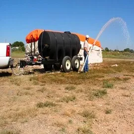 Man using a black water trailer with a spray hose, connected to a truck, to water a dry field in a rural setting.