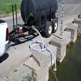 Black water trailer connected to a truck, using a suction hose to draw water from a pond near a stone retaining wall.